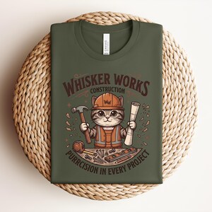 Puede incluir: Camiseta verde oliva con un gato de dibujos animados con ropa de trabajador de la construcci&oacute;n. El gato sostiene un martillo y un plano, con el texto "Whisker Works Construction" y "Purrcision in Every Project". El dise&ntilde;o incluye herramientas.