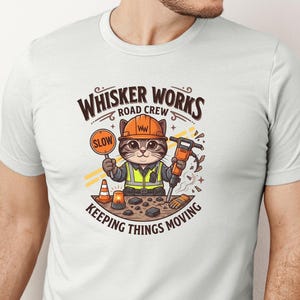 May include: White t-shirt with a cartoon cat dressed as a construction worker. The cat holds a "SLOW" sign and a jackhammer. The text on the shirt reads "Whisker Works Road Crew" and "Keeping Things Moving".