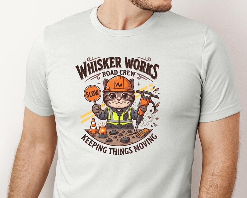 May include: White t-shirt with a cartoon cat dressed as a construction worker. The cat holds a "SLOW" sign and a jackhammer. The text on the shirt reads "Whisker Works Road Crew" and "Keeping Things Moving".