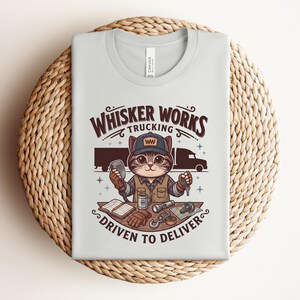 May include: A light grey t-shirt featuring the text "Whisker Works Trucking" and a cartoon cat in a trucker hat. The cat is holding a CB radio and keys. The phrase "Driven to Deliver" is also printed on the shirt.