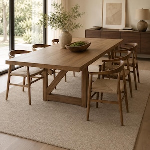 May include: A rectangular wooden dining table with a trestle base, surrounded by six wooden chairs featuring curved backrests and woven seats. A ceramic vase with green foliage and a wooden bowl with moss sit on the table surface.