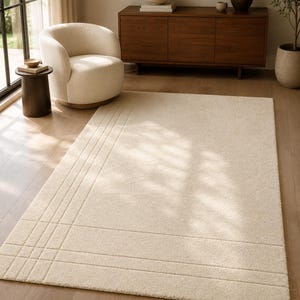 May include: A rectangular area rug in a solid cream color featuring a subtle, raised linear pattern that forms a grid-like design on one side. The rug has a thick, textured pile and is positioned flat on a light-colored wooden floor surface.