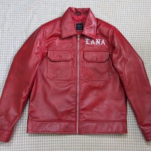 Könnte beinhalten: Eine rote Jacke mit durchgehendem silbernem Reißverschluss, spitzem Kragen und langen Ärmeln. Die Vorderseite hat zwei Brusttaschen mit geknöpften Klappen. Das Wort LANA ist in weißer gotischer Schrift über der rechten Brusttasche gestickt.