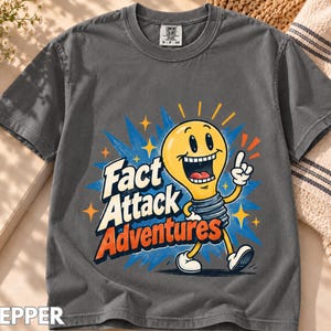 Puede incluir: Una camiseta de manga corta gris carbón con un gráfico de una bombilla amarilla sonriente con brazos y piernas. El texto "Fact Attack Adventures" está impreso en blanco y naranja. La camiseta tiene cuello redondo y una etiqueta.