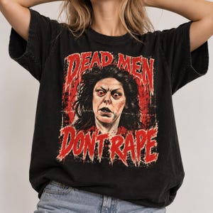 Op de afbeelding: Een zwart t-shirt met korte mouwen en een grafische print van een persoon met donker haar en wijd opengesperde ogen. De tekst "DEAD MEN" staat in het rood boven het portret en "DONT RAPE" in het rood eronder, in een verweerd, druipend lettertype.