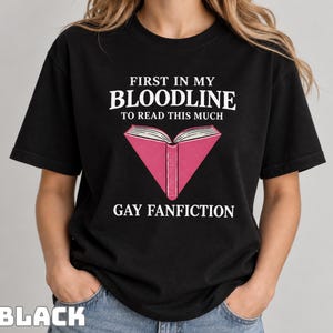 Może przedstawiać: Czarny t-shirt z krótkim rękawem z białym napisem FIRST IN MY BLOODLINE TO READ THIS MUCH GAY FANFICTION, wyśrodkowanym nad i pod grafiką przedstawiającą otwartą różową książkę z białymi stronami i ciemnoróżowym grzbietem.