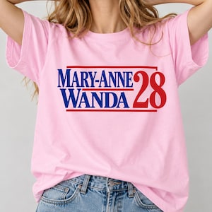 May include: A light pink short-sleeved t-shirt featuring a graphic print in blue and red. The text reads MARY-ANNE WANDA in blue, stacked above each other, with the number 28 in large red font to the right, all enclosed by red horizontal lines.