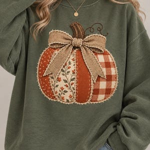 Pode incluir: Uma camisola de gola redonda verde-azeitona com uma grande aplicação de abóbora ao centro. A abóbora é composta por secções de tecido patchwork em tons de laranja, creme e castanho, incluindo padrões florais e axadrezados, com um laço de juta.