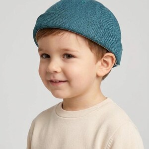 Könnte beinhalten: Eine türkisfarbene Strickmütze. Die Mütze hat eine runde Krone und einen umgeschlagenen Rand. Die Mütze wird von einem kleinen Kind getragen. Das Kind trägt einen beigen Rundhals-Pullover. Der Hintergrund ist einfarbig hellgrau.