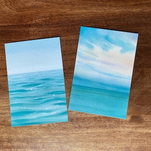 以下が含まれることがあります： 海辺の風景を描いた2つの長方形の水彩画。1つは澄み切った青い空とさざ波立つ水面を描いています。もう1つは雲のある空と海を表す青色のグラデーションが特徴です。どちらも木製の表面にあります。