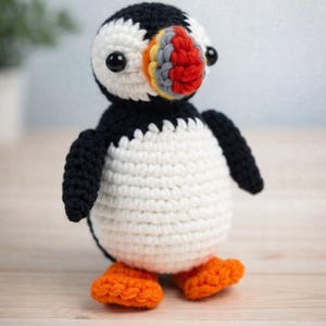 May include: A handmade crochet puffin bird with a black head and wings, a white body, and orange feet. The puffin has a colorful beak with red, yellow, and gray accents. The bird is a whimsical, handcrafted item.