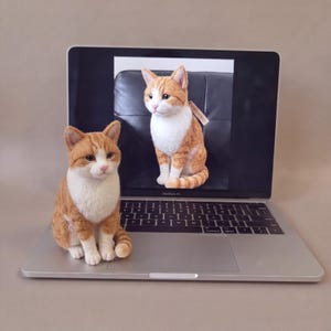 May include: A needle-felted orange and white tabby cat figurine sits on the keyboard of a silver laptop. The laptop screen displays a photograph of the same cat figurine, showing the cat in a seated position with a white chest and paws.