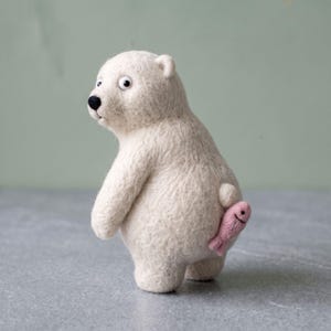 Puede incluir: Una figura de oso polar hecha de lana fieltrada con aguja en color blanco roto, con nariz negra y ojos redondos. El oso está de pie y sostiene un pequeño pez de fieltro rosa claro contra su costado, con una textura difusa en todo el cuerpo.