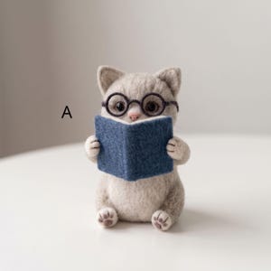 May include: A needle-felted light grey cat figurine with dark brown round glasses, holding a small blue book. The letter A is printed to the left of the cat. The cat has a pink nose and visible paw pads on its feet, sitting in an upright position.