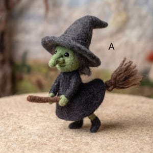 Puede incluir: Una figura de lana fieltrada de una bruja con cara verde, sombrero puntiagudo gris oscuro y vestido gris oscuro. La figura sostiene una escoba marrón con un extremo de cerdas texturizadas. La letra A está impresa en negro arriba a la derecha.