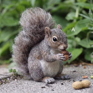 May include: A needle-felted squirrel figurine with textured gray and brown fur, a large bushy tail, and a white chest. The squirrel is holding a small brown acorn in its front paws. The letter A is visible in the upper right corner of the image.