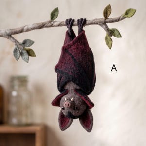 Peut inclure: Une chauve-souris en laine feutrée suspendue à l'envers à une branche feutrée avec des feuilles vertes. La chauve-souris a un corps bordeaux foncé et noir, de petites crocs blancs et un nez rose. La lettre A est imprimée à droite.