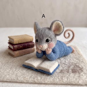 Peut inclure: Une figurine de souris grise en laine feutrée portant un pull bleu, reposant son menton sur ses mains tout en étant allongée. La souris est placée à côté d'une pile de trois petits livres et d'un livre ouvert. La lettre A est visible au-dessus.