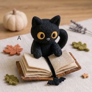 Puede incluir: Un gato negro de lana fieltrada con grandes ojos amarillos sentado tras un libro abierto de páginas beige. Un marcapáginas de cinta de terciopelo negro con un pequeño dije de murciélago cuelga del libro. La letra A es visible a la izquierda.