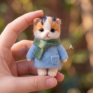 May include: A small needle-felted calico cat figurine featuring a white, orange, and black head, wearing a light blue coat with a small pocket and a green scarf. The letter A is printed in white on the right side of the miniature cat figure.