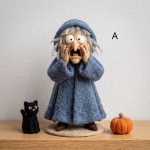 Puede incluir: Una figura de lana fieltrada de una persona con cabello gris que viste un abrigo azul con capucha, con las manos presionadas contra su rostro en expresión de sorpresa. Junto a la figura hay un pequeño gato negro y una calabaza naranja. La letra A es visible.