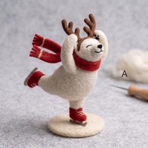 Op de afbeelding: Een naaldgevilte witte berenfiguur met bruin rendiergewei en een rode sjaal, in een schaatshouding op een ronde basis. De beer draagt rode schaatsen met zilverkleurige ijzers en de letter A is rechts van de figuur zichtbaar.