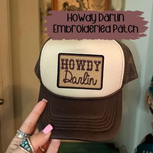 May include: A brown and tan trucker hat with a rectangular embroidered patch that reads "Howdy Darlin". The hat has a brown brim and mesh back, with a tan front panel. The patch is brown with tan lettering.