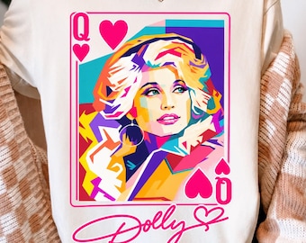 Puede incluir: Una camiseta de color crema con un gráfico pop-art vibrante de una mujer estilizada como una carta de reina de corazones. El diseño incluye el texto "Dolly" en una fuente cursiva rosa debajo del gráfico de la carta, que presenta colores vivos.