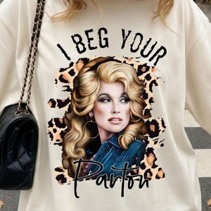 Könnte beinhalten: Ein cremefarbenes T-Shirt mit einem Grafikdruck einer blonden Person mit voluminösem Haar, die eine Jeansjacke und Creolen trägt. Das Design umfasst einen Leopardenmuster-Hintergrund und den schwarzen Text I BEG YOUR Parton.