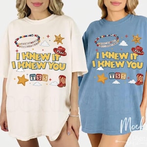 May include: Two short-sleeve t-shirts, one cream and one blue, featuring a graphic print with the text "I KNEW IT I KNEW YOU" and "TS5". The design includes a beaded bracelet, a sheriff badge, a cowboy hat, a star, clouds, and a cowboy boot.
