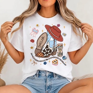 May include: A white t-shirt featuring a graphic print of a brown cowboy hat, a cow-print boot with the name TAYLOR and the number 13, stacked alphabet blocks, a beaded string, a bow, and small floral accents scattered around the central design.