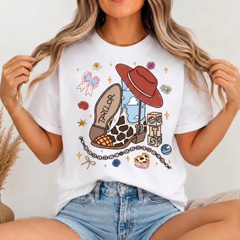 May include: A white t-shirt featuring a graphic print of a brown cowboy hat, a cow-print boot with the name TAYLOR and the number 13, stacked alphabet blocks, a beaded string, a bow, and small floral accents scattered around the central design.
