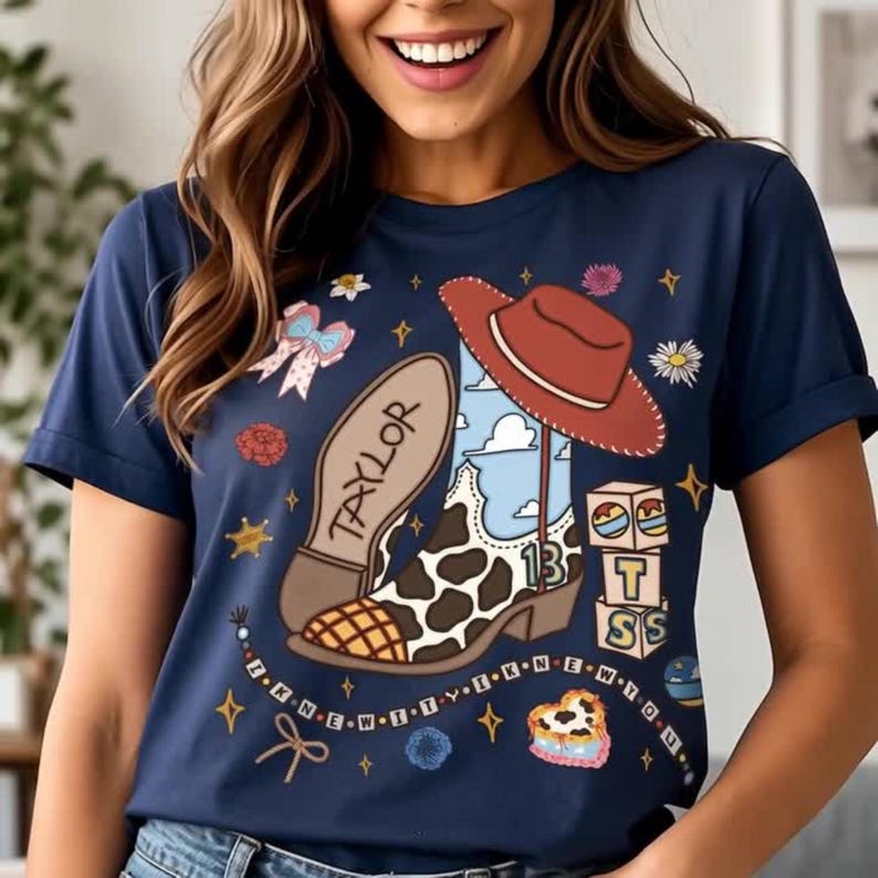 May include: A navy blue t-shirt featuring a graphic print of a cow-print cowboy boot with the name Taylor on the sole, a red cowboy hat, a stack of blocks with the letters T, S, and the number 13, a bow, flowers, and a beaded string with text.
