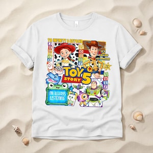 May include: A white short-sleeved t-shirt featuring a colorful graphic print of Toy Story characters including Jessie, Woody, and Buzz Lightyear. The design includes the text TO INFINITY & BEYOND, TOY STORY 5, IT'S CALLED A PONCHO, and I'M ALWAYS LISTENING.