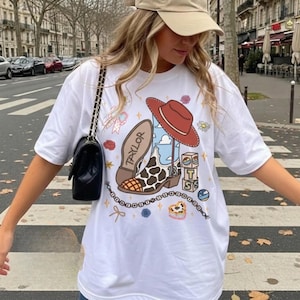 May include: A white t-shirt featuring a graphic print of a brown and white cow-print cowboy boot with the name Taylor on the sole, a red cowboy hat, stacked blocks with the letters T, S, and a number 13, a beaded chain, and small floral accents.