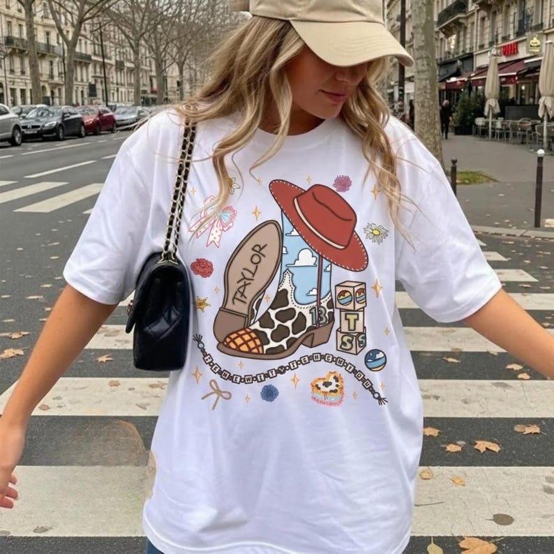 May include: A white t-shirt featuring a graphic print of a brown and white cow-print cowboy boot with the name Taylor on the sole, a red cowboy hat, stacked blocks with the letters T, S, and a number 13, a beaded chain, and small floral accents.