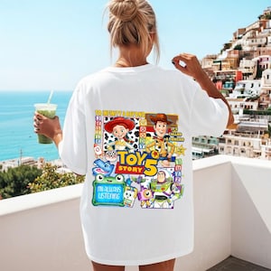 May include: A white t-shirt featuring a colorful graphic on the back with Toy Story characters including Jessie, Woody, and Buzz Lightyear. The design includes the text To Infinity & Beyond, Toy Story 5, and I'm Always Listening, along with various icons.