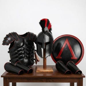 May include: A black and red armor set including a muscle cuirass, a Corinthian-style helmet with a black and red crest, a round shield featuring a red Lambda symbol, and four greaves. The pieces are displayed together as a complete set.