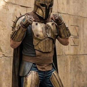 May include: A full-body suit of armor featuring a metallic bronze helmet with a T-shaped visor and spikes, matching shoulder pauldrons, gauntlets, and chest plate. The armor is worn over a dark shirt, blue jeans, and a long brown cape.