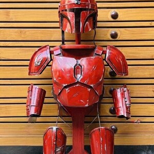 May include: A red Mandalorian armor set, including a helmet, chest plate, shoulder pads, gauntlets, and leg armor, displayed on a wooden stand. The armor has a distressed finish, suggesting battle wear.