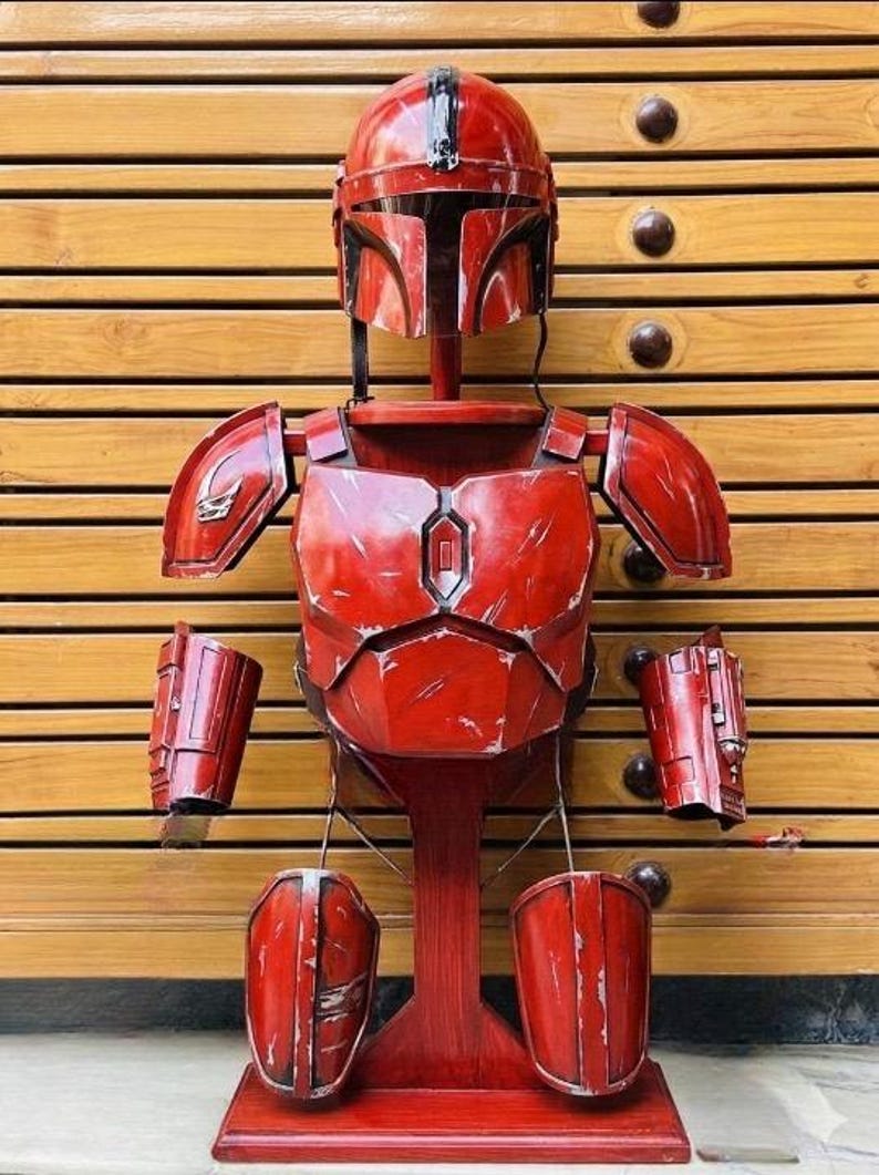 May include: A red Mandalorian armor set, including a helmet, chest plate, shoulder pads, gauntlets, and leg armor, displayed on a wooden stand. The armor has a distressed finish, suggesting battle wear.