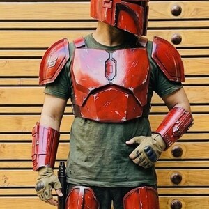 May include: A red Mandalorian helmet and armor set. The costume includes a helmet, chest plate, shoulder pads, arm guards, and knee pads. The armor is weathered with a metallic finish. The person is wearing a green shirt and gloves.