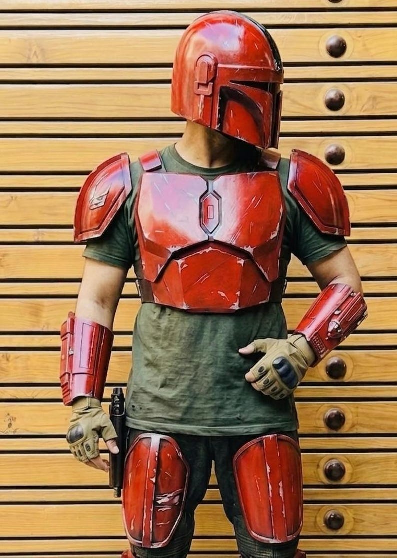 May include: A red Mandalorian helmet and armor set. The costume includes a helmet, chest plate, shoulder pads, arm guards, and knee pads. The armor is weathered with a metallic finish. The person is wearing a green shirt and gloves.
