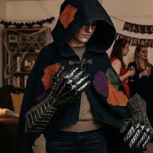 May include: A black hooded cloak featuring irregular orange, purple, and green fabric patches, secured with a toggle closure. The garment is worn with metallic, articulated gauntlets that cover the hands and forearms with segmented plating.