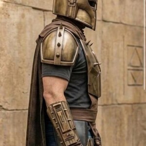 May include: A metallic gold-toned helmet with spikes, matching shoulder pauldrons, and a forearm gauntlet featuring a blue triangular accent. The armor is worn over a dark gray t-shirt and blue jeans, complemented by a long brown cape draped over the back.