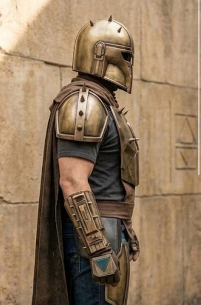 May include: A metallic gold-toned helmet with spikes, matching shoulder pauldrons, and a forearm gauntlet featuring a blue triangular accent. The armor is worn over a dark gray t-shirt and blue jeans, complemented by a long brown cape draped over the back.