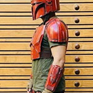 May include: Red Mandalorian armor set, including a helmet, chest plate, shoulder pads, arm guards, and leg armor. The armor is worn over a green shirt and brown belt. The gloves are tan and black.