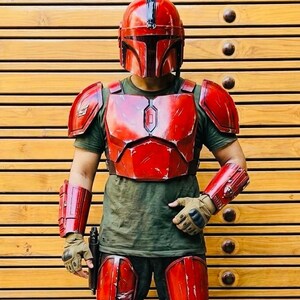 May include: A red Mandalorian armor set, including a helmet, chest plate, shoulder pads, arm guards, and knee pads. The armor is worn over a green shirt and gloves. A black pistol is held in the right hand.