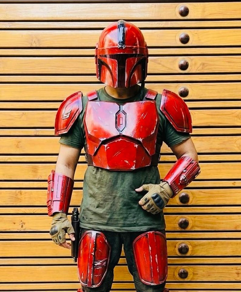 May include: A red Mandalorian armor set, including a helmet, chest plate, shoulder pads, arm guards, and knee pads. The armor is worn over a green shirt and gloves. A black pistol is held in the right hand.