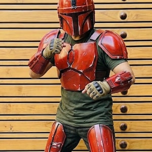 May include: A red Mandalorian armor set, including a helmet, chest plate, shoulder pads, arm guards, and knee pads. The armor has a weathered appearance, suggesting battle. The person is wearing a green shirt and gloves. The background is a wooden wall.